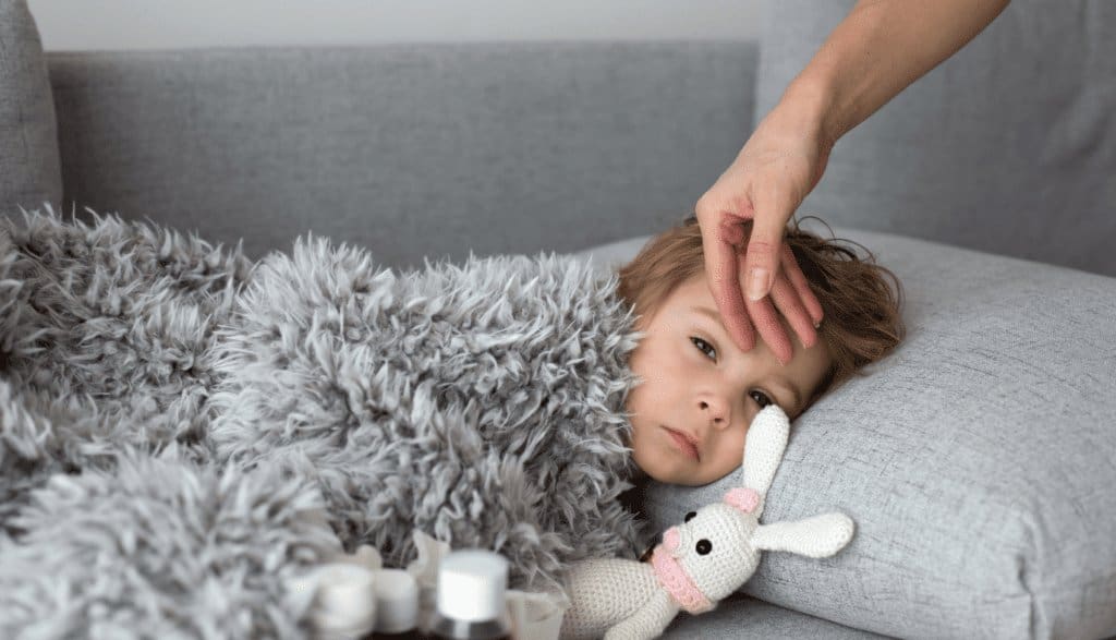 Young child feeling unwell, wrapped in a soft blanket on the couch with a caring hand on their forehead and a comfort toy nearby.